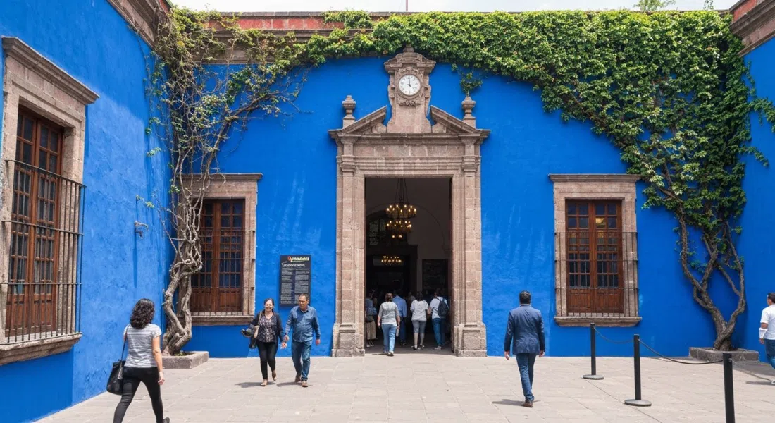 el patio azul y la entrada frondosa del Museo Frida Kahlo, con visitantes entrando bajo el sol brillante del mediodía